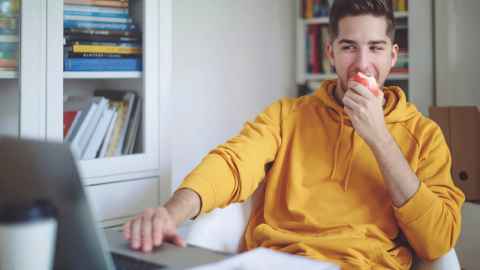 Young man eating apple while at desk and on computer